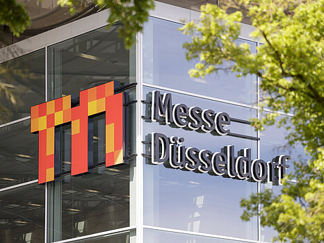 Exteriören på en modern glasbyggnad med logotypen "Messe Düsseldorf" tydligt placerad på fasaden, delvis inramad av gröna trädgrenar.
