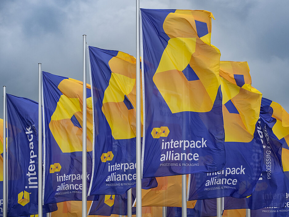 Flera blå flaggor vajar, med den gul-orange logotypen och texten ”interpack alliance – Processing & Packaging” mot en molnig himmel.